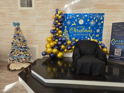 A stage containing a black chair with a blue Christmas banner with a balloon garland and a Christmas tree to the left of the stage
