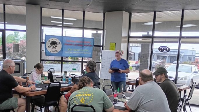 5.28.26 Trivia Night A person in a blue shirt stands in front of two tables full of people with a clipboard