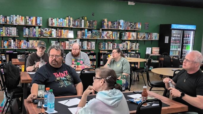 5.28.26 Trivia Night A group of people deep in thought sit around tables in front of a green wall filled with board game shelves