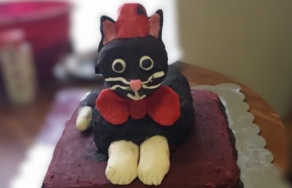A cake shaped like a cartoonish black cat with white paws wearing a red bow tie and red fez