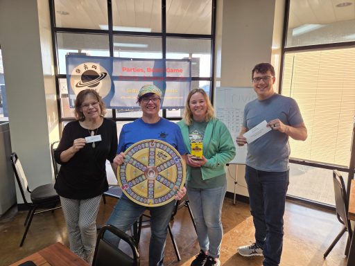 4.23.26 Trivia Night Winners Four people stand in front of a window with a banner. One of them is holding a Trivial Pursuit board.