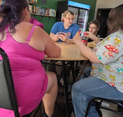 7.1.26 Uno Tournament Four people play Uno at a table in front of a drink cooler and a green wall with shelves of board games.