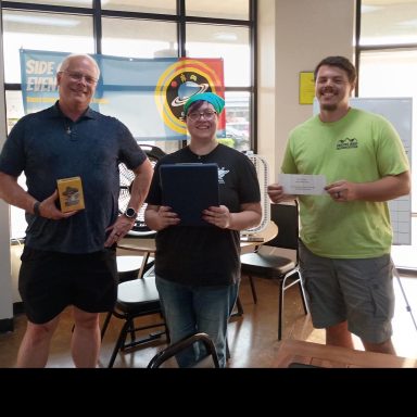 8.6.26 Trivia Night Winners Three people stand in front of a window with a banner. One holds a game, one holds a clipboard, and one holds a gift certificate.