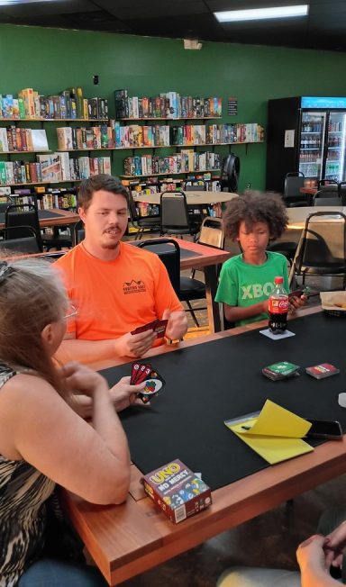 8.5.26 Uno Tournament Three people play Uno at a table in front of a green wall full of board game shelves.
