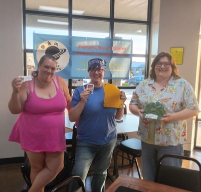 7.1.26 Uno Tournament - Winners Three people stand in front of a banner in a window. Each is holding a prize.
