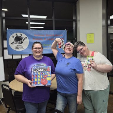 6.3.26 Uno Tournament - Winners Three adults stand in front of a Side Quest Events banner. Two are holding games and the other is holding a hand of Uno cards.