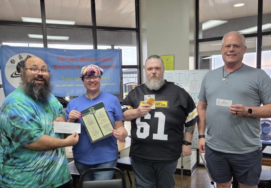 6.11.26 Trivia Night Winners Four people stand in front of windows with a blue banner holding prizes and a menu