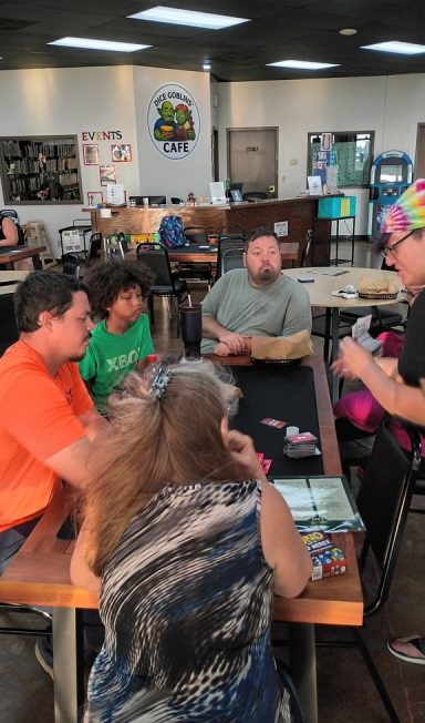 8.5.26 Uno Tournament Four players sit around a table while a fifth person explains a card game.