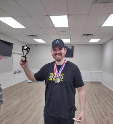 Board Game Olympics Champion A man in a black shirt and black cap with medals around his neck holds up a trophy