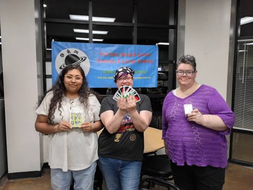 Two people hold board games on either side of a person holding a large hand of Uno cards