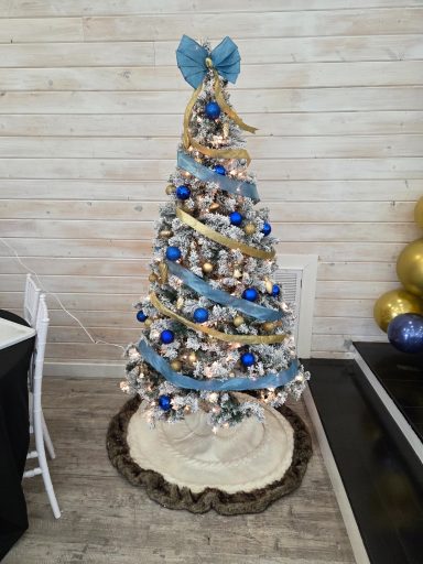 A flocked Christmas tree decorated with blue and gold ornaments and ribbon and topped by a blue and gold bow