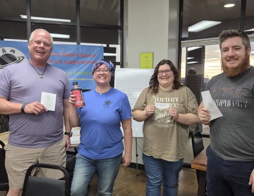 6.25.26 Trivia Night Winners Four people stand in front of a blue banner hanging in a window. Three are holding slips of paper and the fourth is holding a drink bottle.