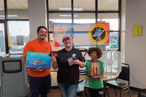 8.5.26 Uno Tournament - Winners Three people stand in front of a banner in a window. Two hold board games and the other holds a hand of Uno cards and the card box.
